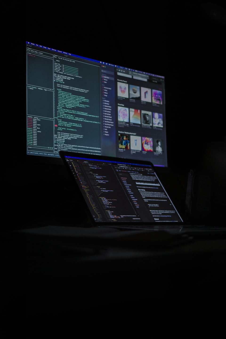 Laptop and monitor displaying complex code in a dark room.